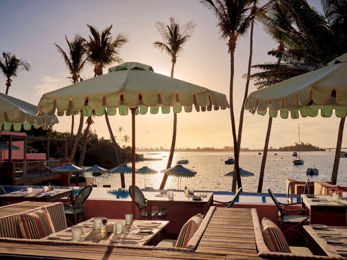 A seaside view at sunset featuring palm trees, beach umbrellas, lounge chairs, and boats on calm water. A serene vacation setting.