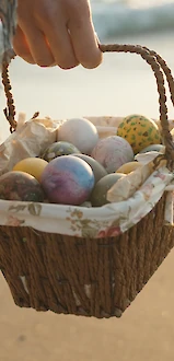 A person holds a woven basket filled with colorful Easter eggs on a sandy beach, wearing a plaid shawl in warm light.