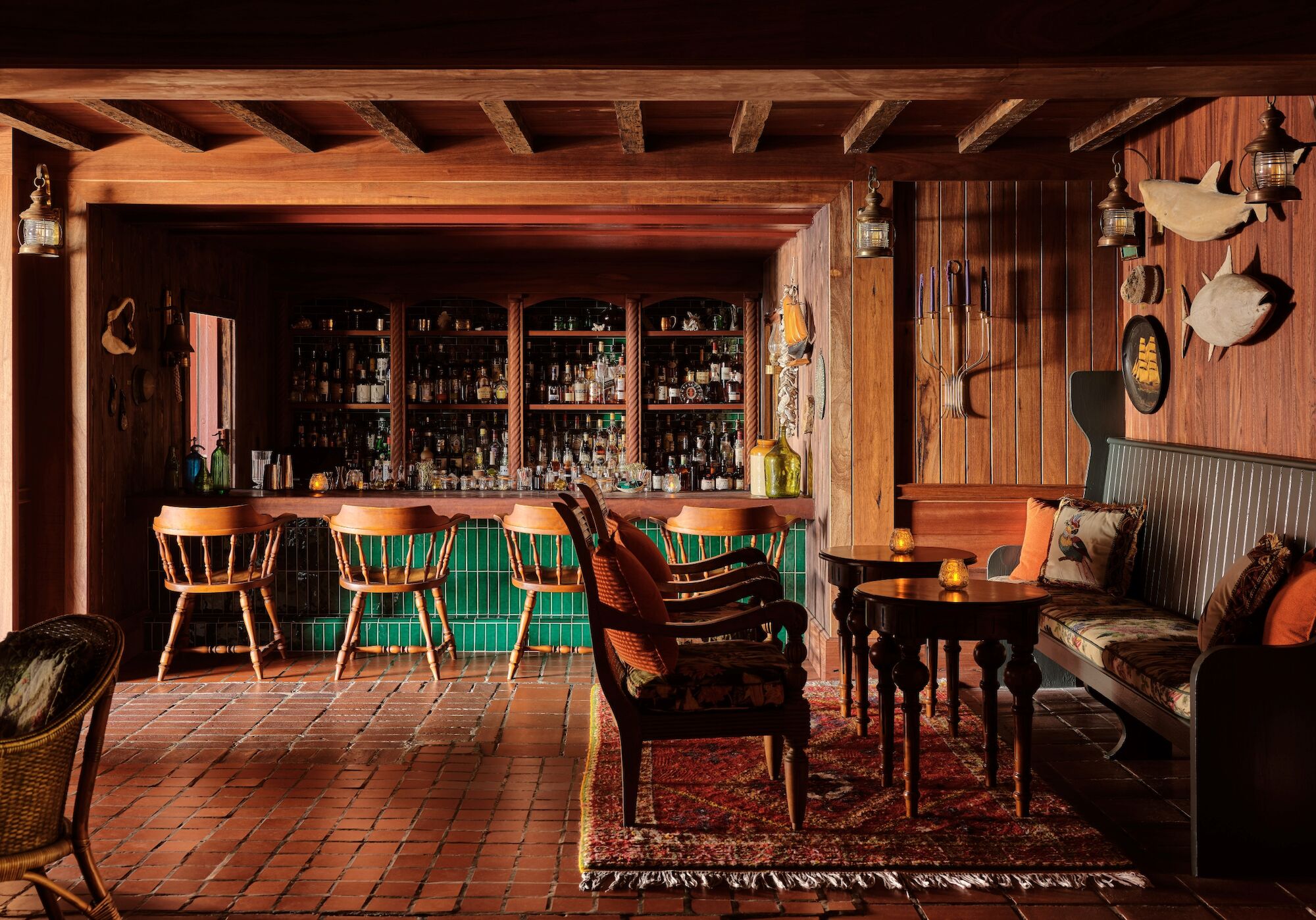 Rustic wooden bar lounge with stocked shelves, green-tiled backdrop, stools and tables, hanging lanterns, and cozy seating.