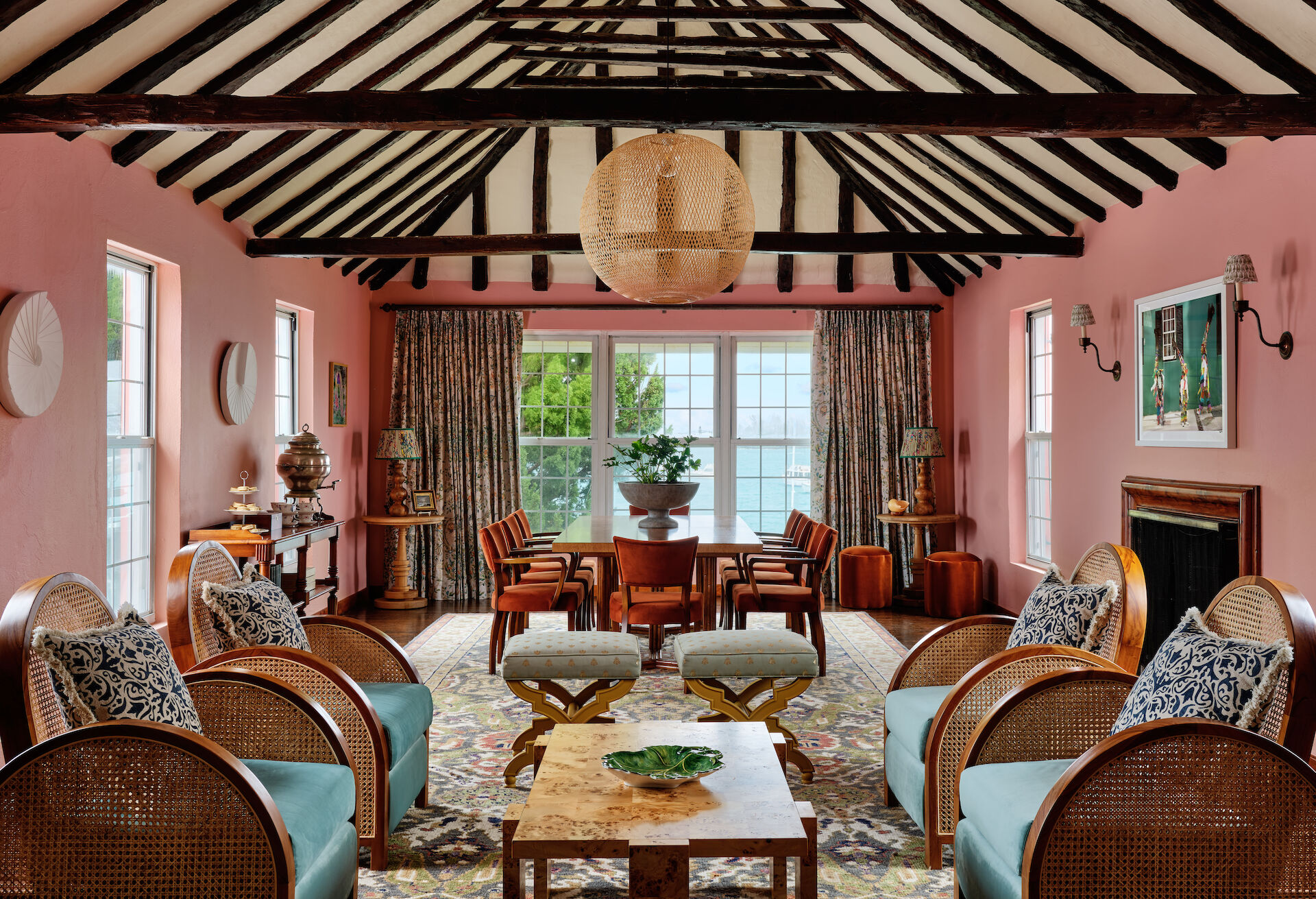 A spacious living room with pink walls, wooden beams, rattan furniture, and large windows adorned with curtains. A dining area sits in the background.