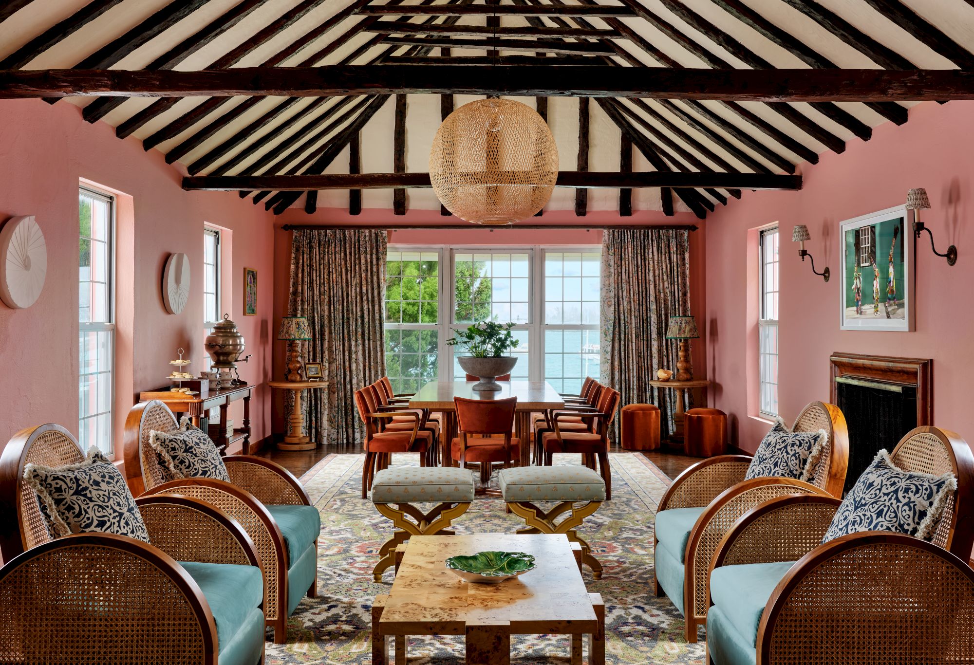A spacious living room with pink walls, wooden beams, rattan furniture, and large windows adorned with curtains. A dining area sits in the background.