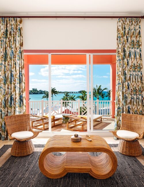 A bright tropical living room with wicker chairs and coffee table, sheer curtains, and a view of palm trees from an open balcony.