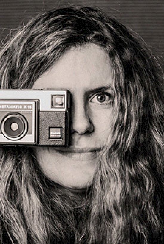A person with wavy hair holds an Instamatic 133 camera up to their eye, creating a playful pose in a black and white photo.