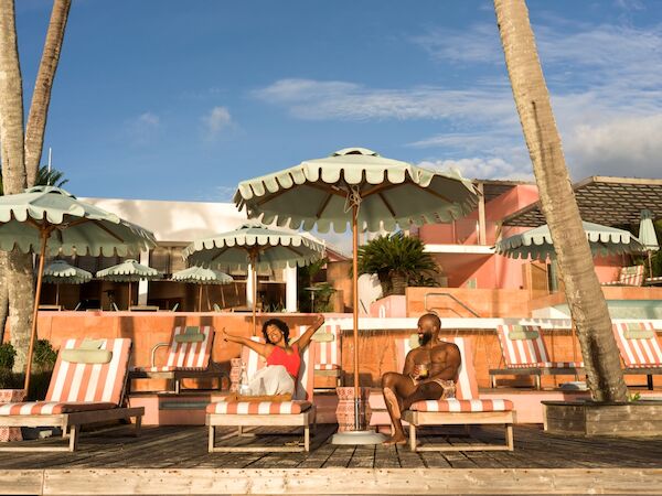 Two people lounge under striped umbrellas by poolside chairs at a sunny beach resort, with palm trees and pastel buildings in the background, smiling.