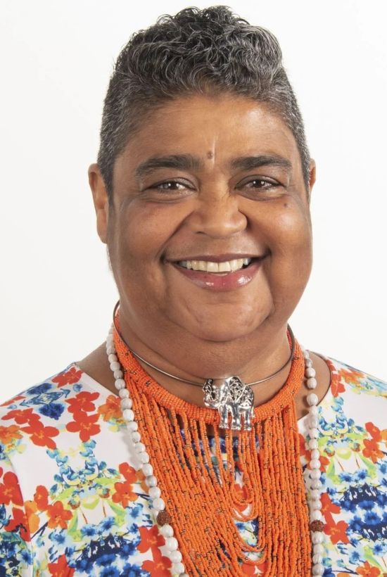 The image shows a person with short hair, wearing a colorful floral patterned top and an orange beaded necklace, smiling in front of a white background.