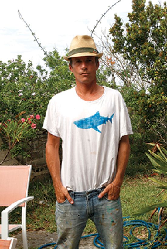 A person stands outdoors wearing a hat and shark graphic T-shirt, hands in pockets, with greenery and outdoor furniture in the background.