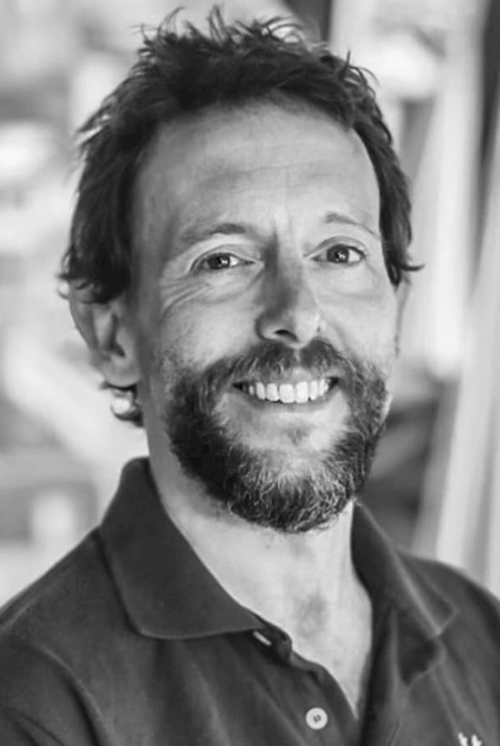 A man with a beard and mustache is smiling at the camera. He is wearing a collared shirt in a casual setting with a blurred background.