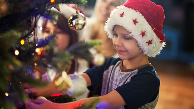 A child in a Santa hat decorates a Christmas tree, with two people in the background. The scene is festive and joyful.