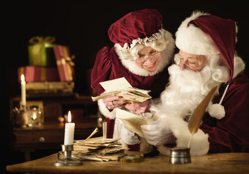Two Santa figures counting money/cards by candlelight, with gifts in the background&mdash;a cozy, festive scene of holiday cheer and exchange.
