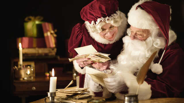 Two Santa figures counting money/cards by candlelight, with gifts in the background&mdash;a cozy, festive scene of holiday cheer and exchange.