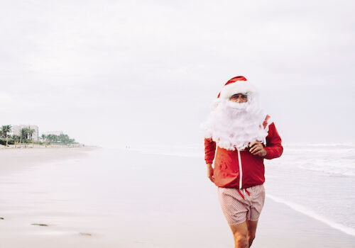 A person dressed as Santa is running on a beach, wearing a red jacket, shorts, and a Santa hat, with palm trees in the background.