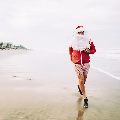 A person dressed as Santa is running on a beach, wearing a red jacket, shorts, and a Santa hat, with palm trees in the background.