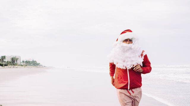 A person dressed as Santa is running on a beach, wearing a red jacket, shorts, and a Santa hat, with palm trees in the background.