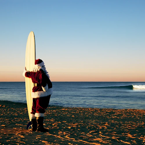 A person dressed as Santa Claus stands on a beach, holding a surfboard, looking at the ocean during a sunset or sunrise.