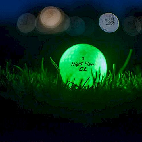 A glowing green golf ball labeled "Night Flyer CL" rests on grass with blurry lights in the background at night.