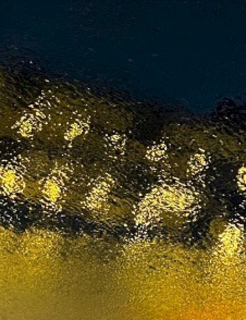 The image shows a blurred view, possibly of lights through a textured or wet surface, creating a shimmering, abstract effect across dark and yellow hues.