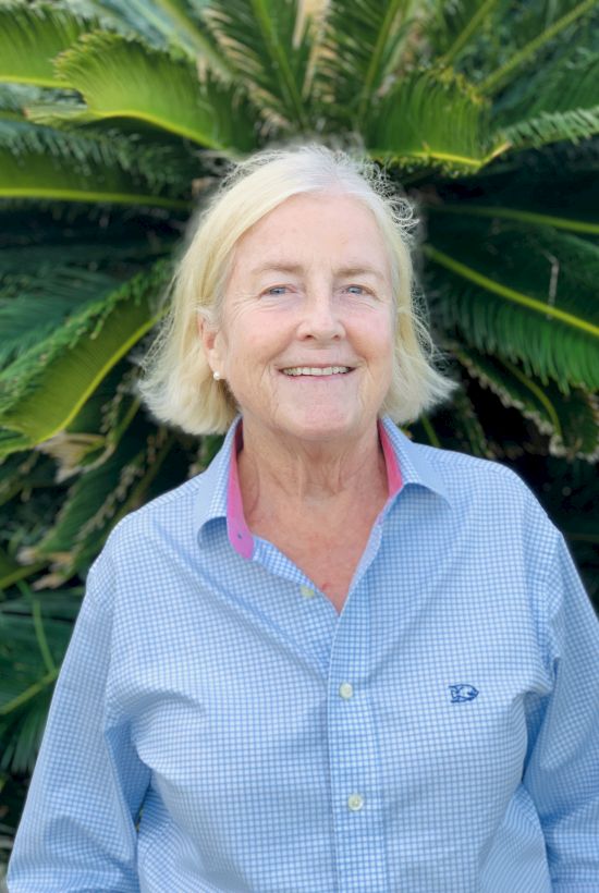 A person with short blonde hair is smiling, wearing a blue shirt, standing in front of large green foliage.