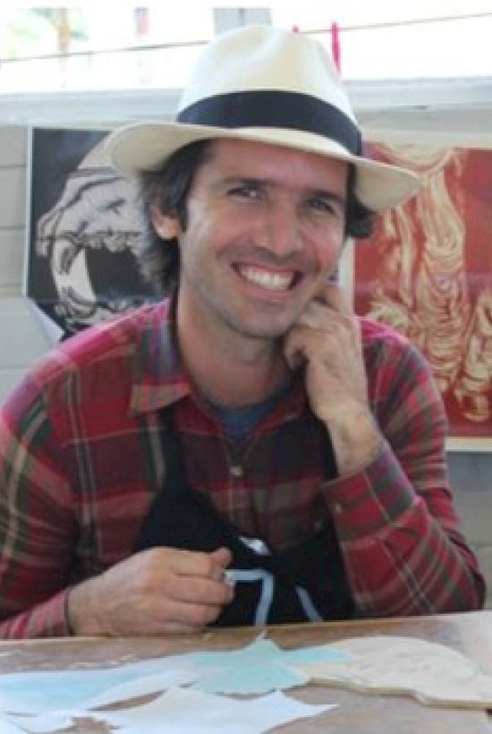 A person wearing a hat and a plaid shirt, smiling while sitting at a table covered with papers and art supplies, with artwork displayed behind.