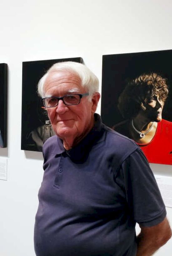 An older man wearing glasses stands in front of portrait paintings in an art gallery, appearing to be enjoying the exhibition.