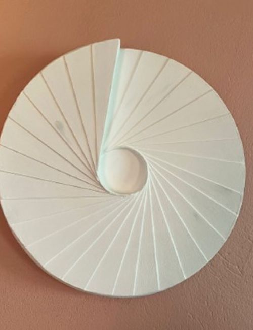 The image shows a white, spiral-shaped wall-mounted artwork with a circular design, displayed against a muted pink background.