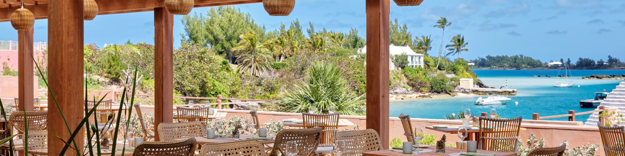 An outdoor seating area with wooden tables and wicker chairs under a pergola, overlooking a scenic coastal view with clear blue water and greenery.