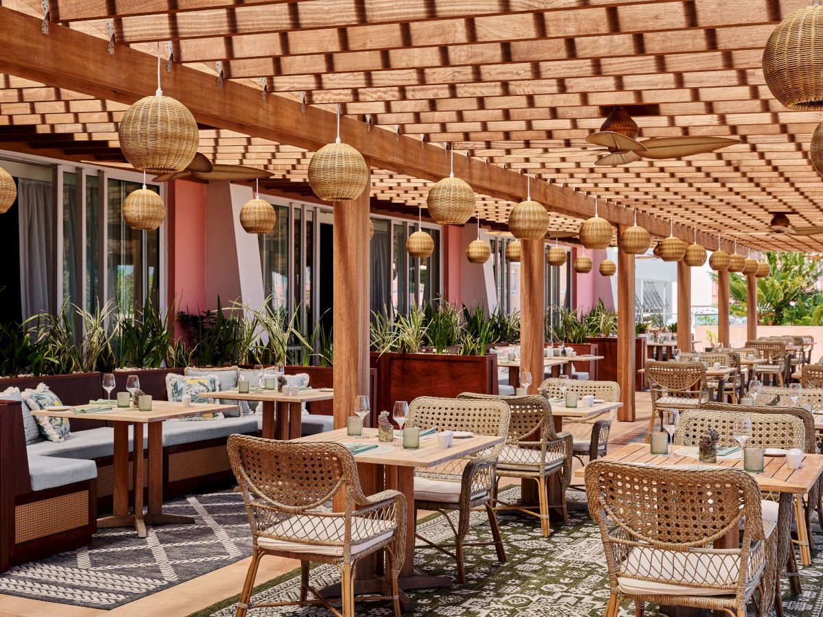 An outdoor restaurant with woven furniture, hanging lights, and a wooden pergola, featuring a tropical ambiance with numerous tables set for dining.