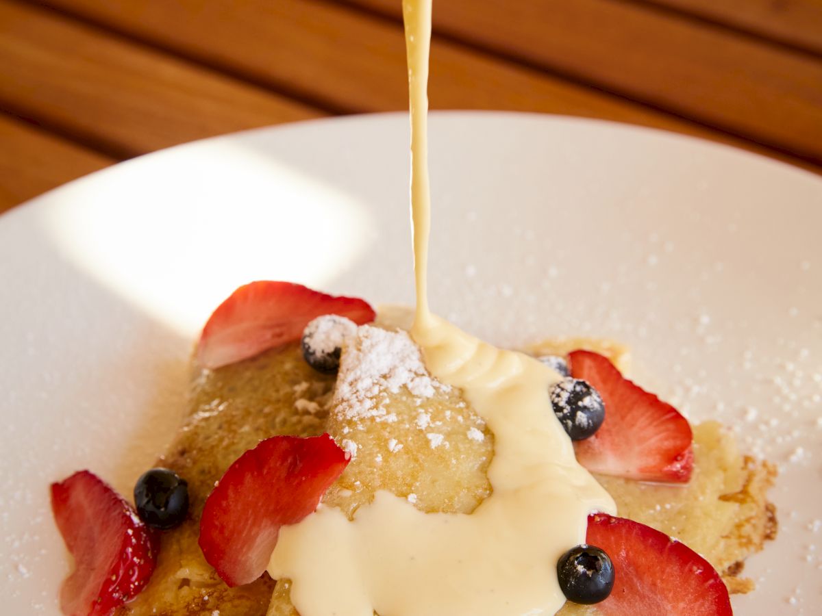 A person drizzling a creamy sauce over a dessert garnished with strawberries, blueberries, and powdered sugar, served on a white plate.