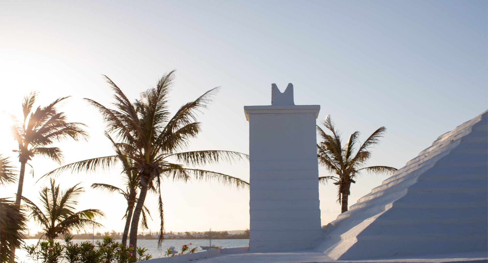 A white pillar and stairs by the sea at sunset, framed by tall palm trees and a clear sky.
