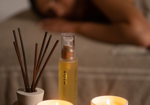 Lit VOYA candles and a reed diffuser on a wooden tray, with a blurred person resting in the background, glowing warmly.