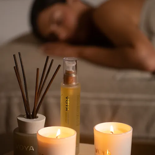 Lit VOYA candles and a reed diffuser on a wooden tray, with a blurred person resting in the background, glowing warmly.