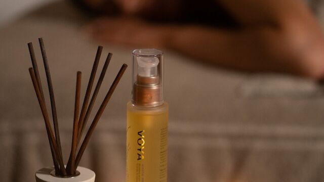 Lit VOYA candles and a reed diffuser on a wooden tray, with a blurred person resting in the background, glowing warmly.
