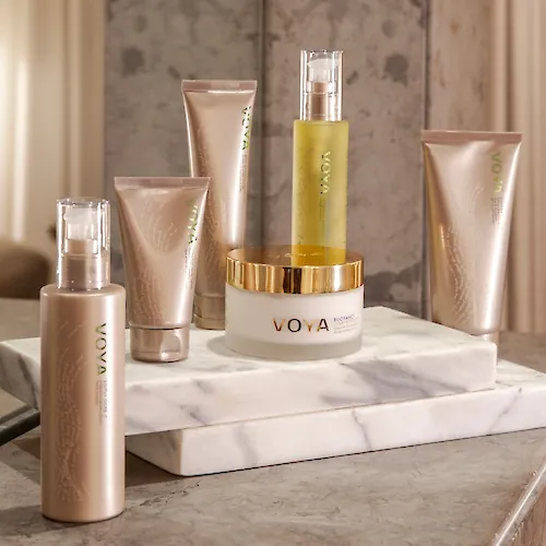 A set of VOYA skincare bottles and tubes on a marble pedestal in a bathroom setting.