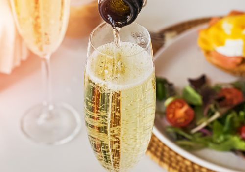 A bottle is pouring sparkling wine into a glass. There are also other glasses and a plate with a salad and an egg dish in the background.