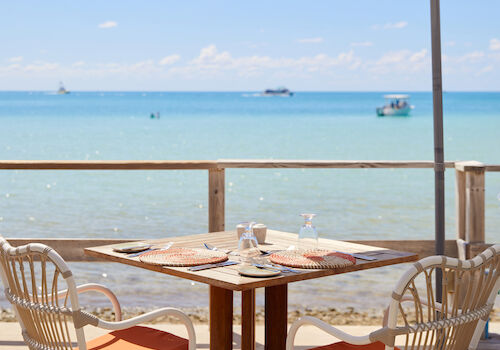 Beachside table with chairs under an umbrella, set for dining with an ocean view and boats in the distance.