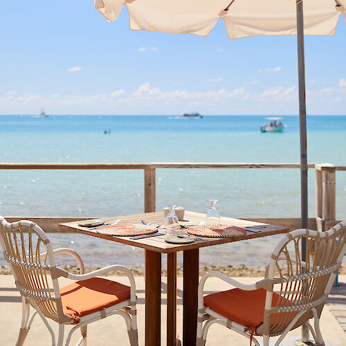 Beachside table with chairs under an umbrella, set for dining with an ocean view and boats in the distance.