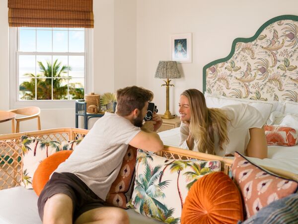 A couple relaxes on a cozy patterned bed as a man takes a photo with a camera and the woman smiles, in a bright room.