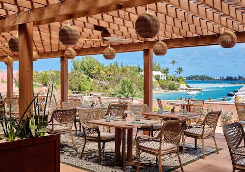 An outdoor dining area with wooden furniture and hanging lights overlooks a scenic ocean view, with lush greenery and clear skies.