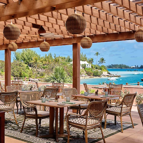 An outdoor dining area with wooden furniture and hanging lights overlooks a scenic ocean view, with lush greenery and clear skies.