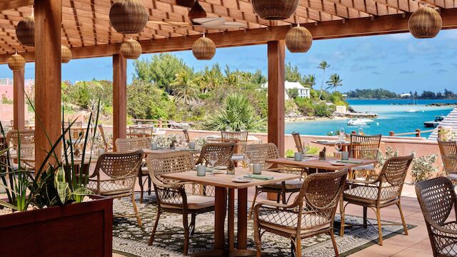An outdoor dining area with wooden furniture and hanging lights overlooks a scenic ocean view, with lush greenery and clear skies.