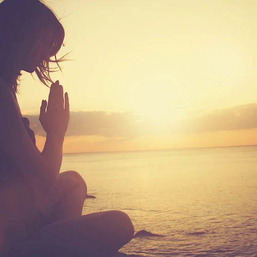 A person sitting by the ocean at sunset, hands together in a meditative or prayer pose, with the sun casting a warm glow over the scene.
