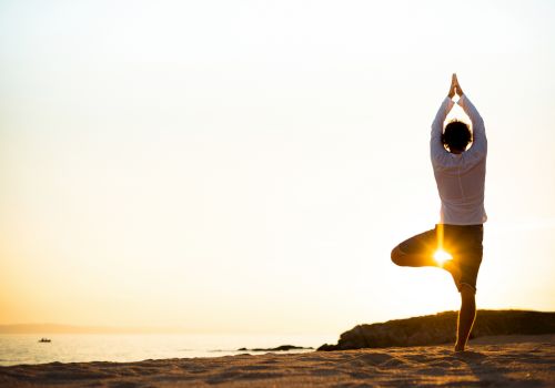A person is doing a yoga pose on the beach at sunset, with the sun shining through their legs, creating a serene and peaceful setting.