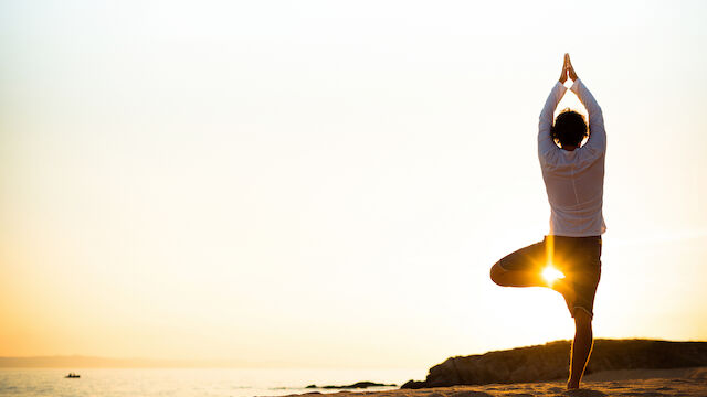A person is doing a yoga pose on the beach at sunset, with the sun shining through their legs, creating a serene and peaceful setting.