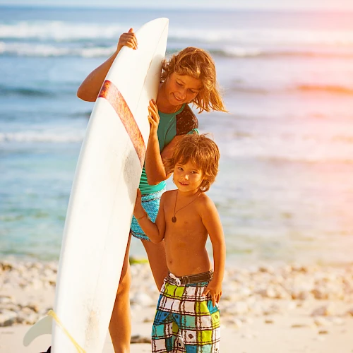 A woman and a young boy stand on a sandy beach with a surfboard, the ocean and sky in the background.