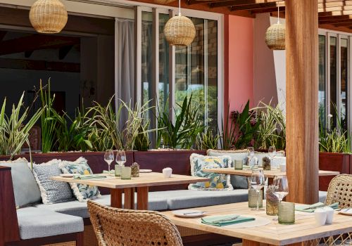The image shows an outdoor dining area with wooden furniture, cushioned seats, hanging wicker lamps, and plants, creating a cozy and inviting ambiance.