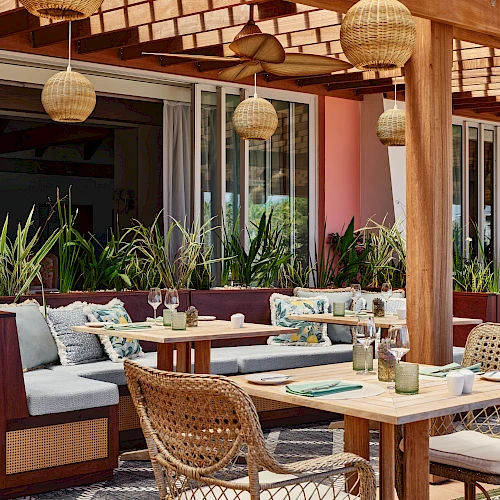 The image shows an outdoor dining area with wooden furniture, cushioned seats, hanging wicker lamps, and plants, creating a cozy and inviting ambiance.