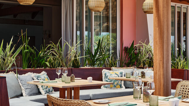 The image shows an outdoor dining area with wooden furniture, cushioned seats, hanging wicker lamps, and plants, creating a cozy and inviting ambiance.