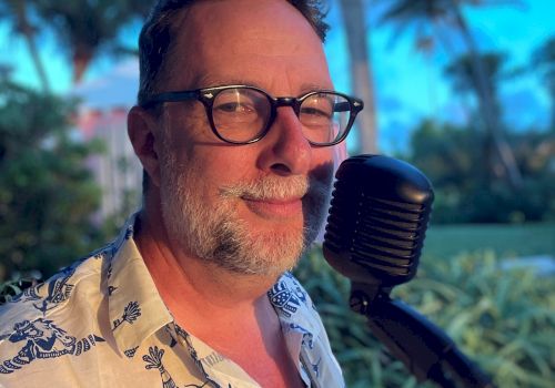 A person wearing glasses and a patterned shirt is standing outdoors with a microphone, with palm trees and a clear sky in the background.