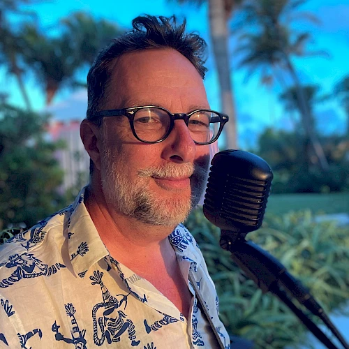 A person wearing glasses and a patterned shirt is standing outdoors with a microphone, with palm trees and a clear sky in the background.