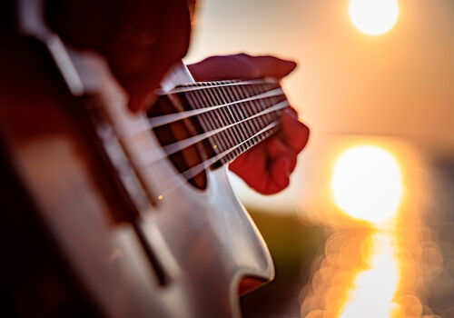 A close-up of a guitar being played, strings and fretboard in focus, with a warm sunset glow reflecting off the water in the background.