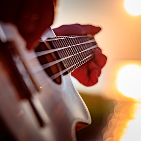 A close-up of a guitar being played, strings and fretboard in focus, with a warm sunset glow reflecting off the water in the background.